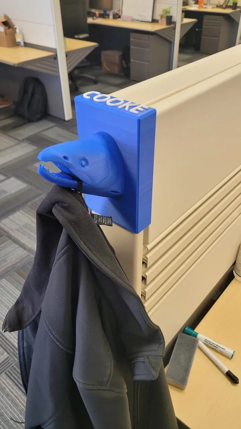 A blue 3D-printed salmon-head coat hook on a nameplate, mounted on an office cubicle partition and holding a jacket