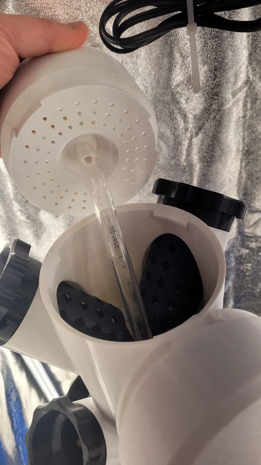 A printed perforated drip-distributor cap held in hand, beside the open tower top and its pour tube